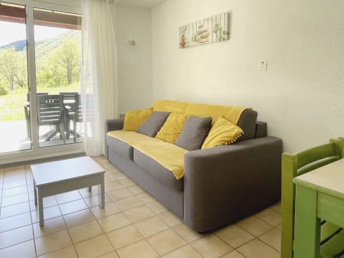 Salon lumineux avec canapé gris, coussins jaunes, vue montagne et accès à la terrasse au Vallon des Sources
