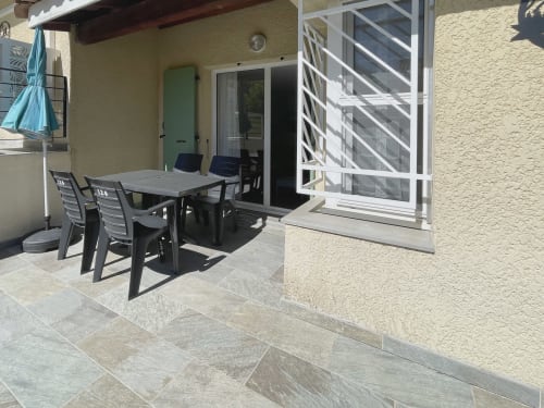 Terrasse ensoleillée au Vallon des Sources, Digne-les-Bains : table de repas avec chaises noires, balustrade blanche, porte verte, pierre