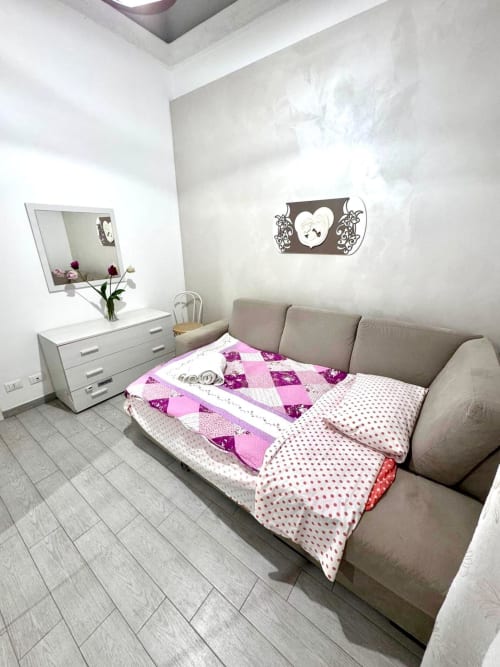 Bright bedroom with beige sofa bed, pink and purple bedding, white dresser with mirror, and decorative wall clock at Casa