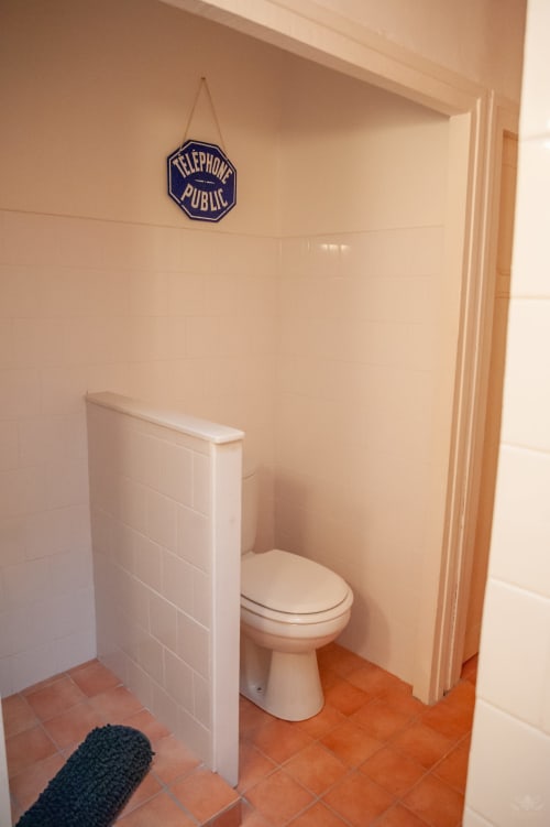 Salle de bains de cottage avec toilettes blanches, murs carrelés, sol en terre cuite et enseigne vintage de téléphone public à Mi Casa Es Su Casa