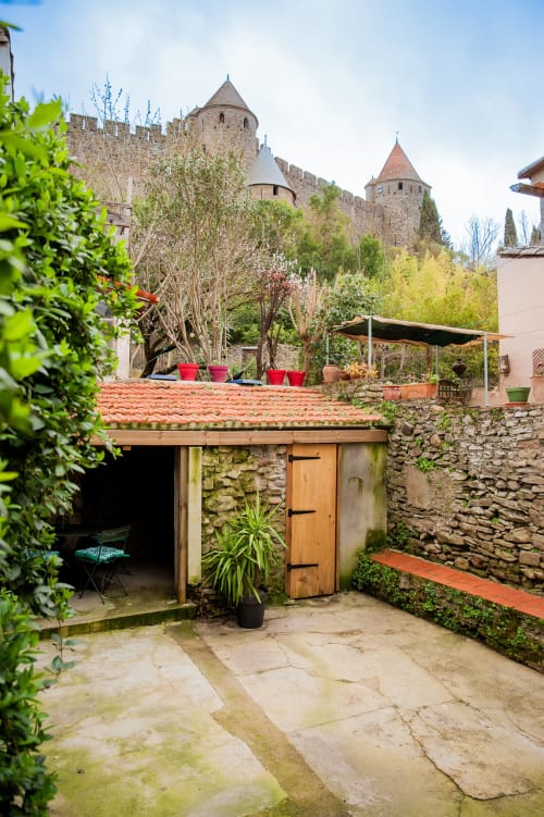 Terrasse de maison en pierre avec les tours de la forteresse de Carcassonne, toiture en tuiles rouges, plantes et vue sur le château médiéval