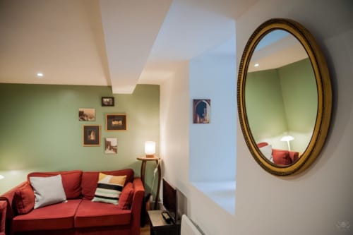 Salon moderne avec canapé rouge, murs verts, miroir ovale orné et tableaux à La Maison de Marcelo, cottage à Carcassonne