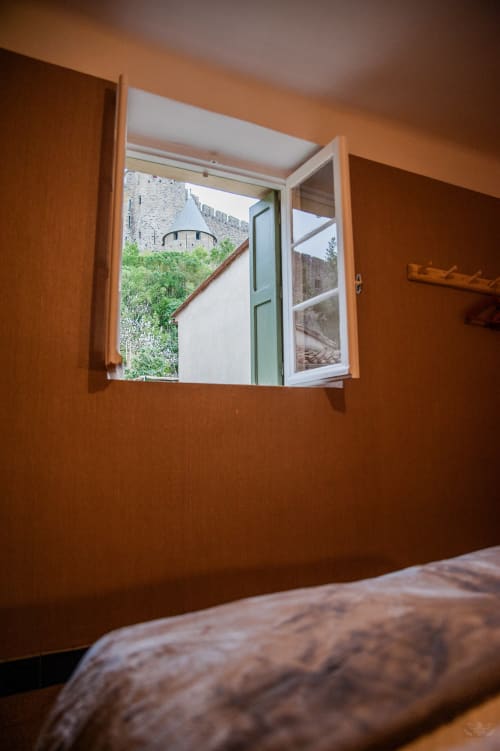Chambre chaleureuse aux tons orangés avec fenêtre ouverte offrant une vue sur le château et les remparts de Carcassonne, gîte La Maison de Marcelo