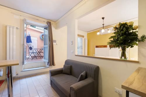 Bright studio room at Casa Scola Loano with gray sofa, yellow accent wall, fresh flowers, and access to pink balcony in Loano