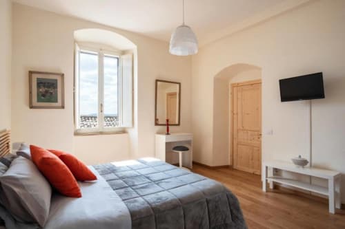 Camera luminosa nella Proprietà Clara ad Assisi con letto, cuscini rossi, vista dalla finestra, TV e porte in legno
