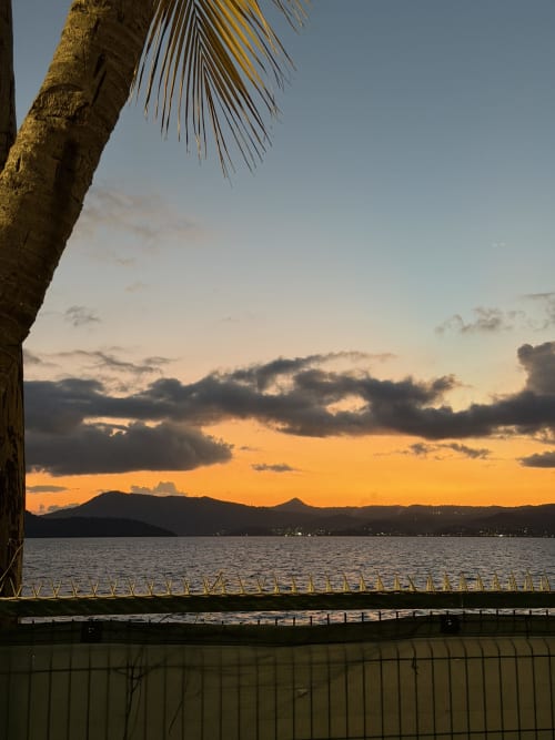 Vue du coucher de soleil depuis la terrasse de Kaz Kafe surplombant la baie de Mamoudzou avec palmiers et montagnes silhouettées sur ciel doré