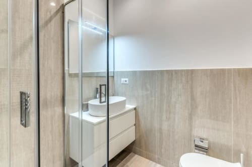 Bagno moderno con lavabo rotondo bianco, piastrelle effetto legno beige, box doccia in vetro e vanità bianca nel centro città