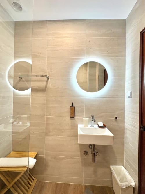 Banheiro moderno da Hospedaria Buganvília Alvor com espelhos circulares iluminados, pia branca e azulejos bege nas paredes