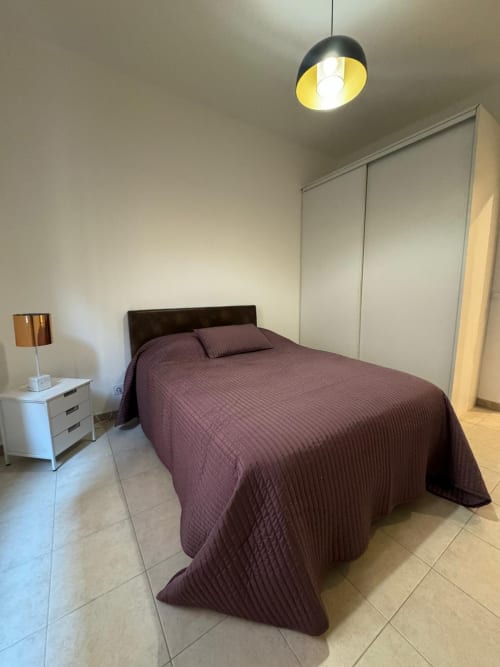 Chambre moderne à l'appartement Altezza à Bastia avec literie violette, table de nuit blanche, lampe suspendue et armoire intégrée.