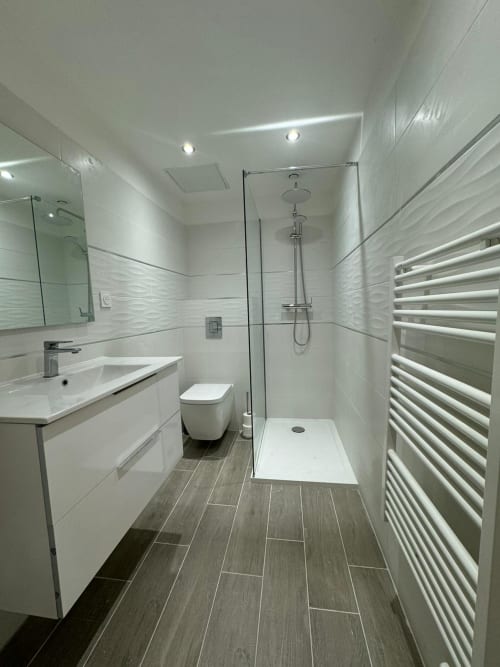 Salle de bain moderne dans un appartement bastiais avec vasque blanche, douche vitrée, porte-serviettes chauffant et sol imitation bois