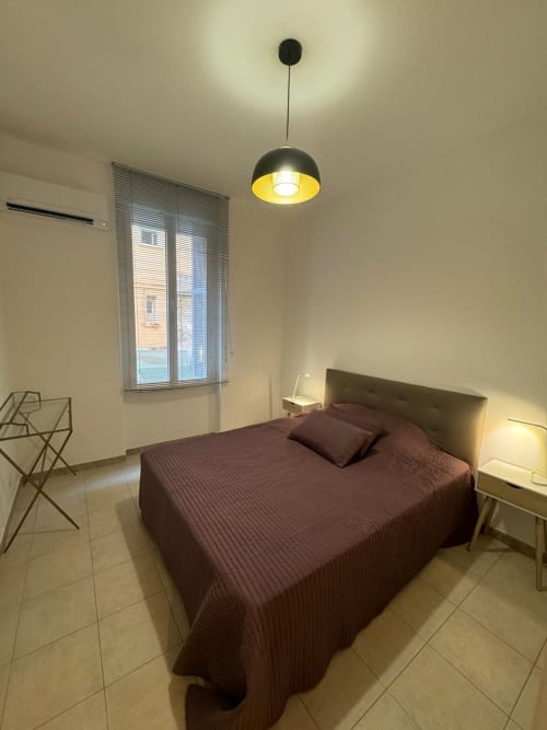 Chambre moderne dans un appartement de Bastia avec lit bordeaux, luminaire suspendu, fenêtre à stores et mobilier minimaliste