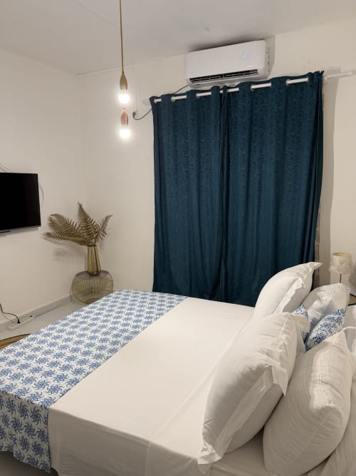 Chambre douillette au Kaz Kafe B&B à Mamoudzou avec lit blanc, rideaux bleu marine, TV et vue sur le jardin