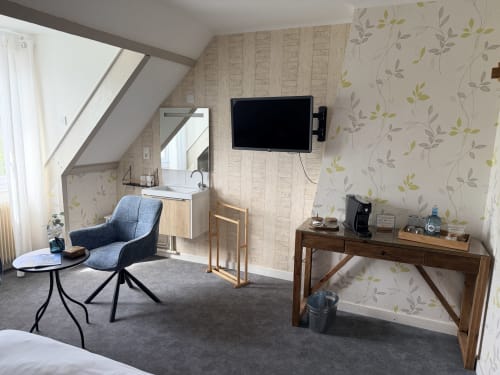 Chambre mansardée moderne aux Vieilles Digues avec fauteuil bleu, bureau, TV murale et vue sur le jardin à Beauvoir