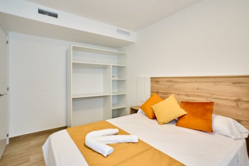 Dormitorio moderno en Tr3sor Homes, Calella, con cama de madera, detalles naranjas, ropa de cama blanca y almacenaje integrado