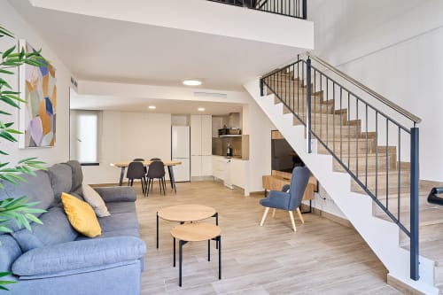 Sala de estar abierta y moderna con sofá azul, comedor, cocina y escalera blanca en apartamento Tr3sor 31, Calella