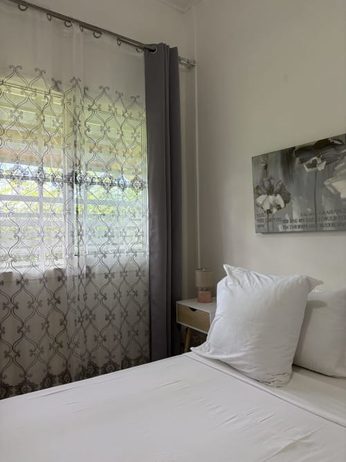 Chambre Zen Bambou du Kaz Kafe avec coussins blancs, rideaux à motifs, table de nuit et vue sur le jardin à Mamoudzou
