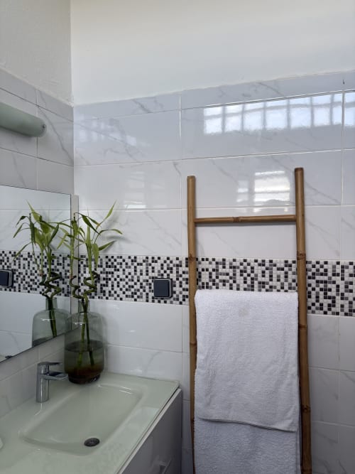 Salle de bains moderne du Kaz Kafe BnB avec carrelage blanc, frise mosaïque noire, échelle porte-serviettes en bambou et plante verte à Mamoudzou