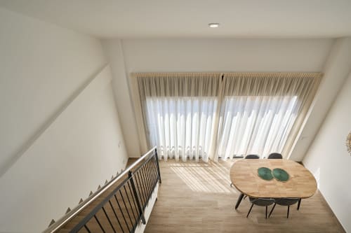 Luminoso comedor dúplex con mesa de madera, escalera y cortinas de piso a techo con vistas a Calella en Tr3sor Homes