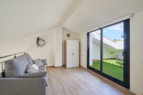 Moderno dúplex con salón luminoso, sofá gris, armario blanco y amplias puertas correderas hacia terraza verde en Calella