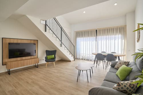 Sala de estar moderna en dúplex de Tr3sor Homes Calella con escalera, sofá gris, comedor, TV y ventanas luminosas