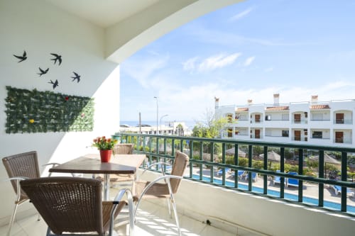 Varanda ensolarada do hotel RB Algarve em Albufeira com assentos, vegetação, decoração com pássaros, vista da piscina e edifícios brancos