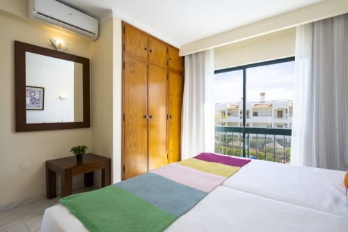 Quarto luminoso do apartamento RB Algarve Albufeira com cama colorida, roupeiro em madeira, espelho e varanda com vista para a piscina