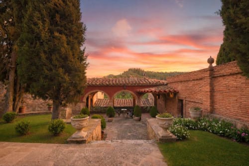 Patio exterior con arcos de ladrillo, cipreses y muros de piedra al atardecer.
