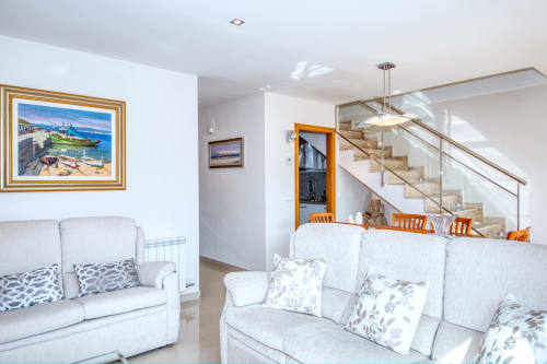 Modern living room with cream sofas, ocean artwork, and glass staircase in a beachfront vacation rental in Costa brava region
