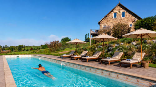 La piscine du gîte Les-instants-volés avec un baigneur dans une eau cristalline, maison en pierre et transats à proximité