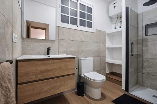 Ba
tiene vanidad de madera, paredes con azulejos grises, inodoro blanco y ducha abierta