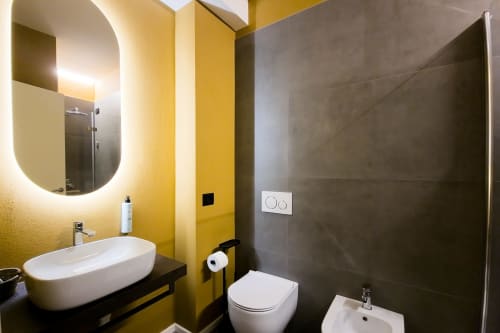 Bagno moderno presso SMalL Guesthouse Bussolengo con pareti gialle, specchio illuminato, lavabo bianco e doccia con piastrelle scure