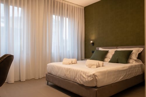 Camera da letto moderna presso SMalL Guesthouse Bussolengo con parete accento verde oliva, biancheria bianca e tende semitrasparenti