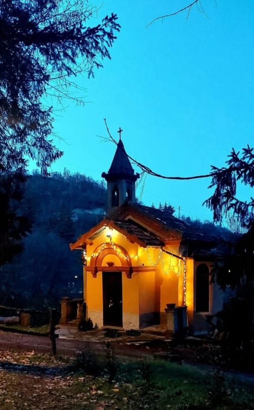 Chapelle jaune illuminée avec croix au crépuscule en Piémont, Italie, entourée d'arbres et de montagnes au Palazzo Losio