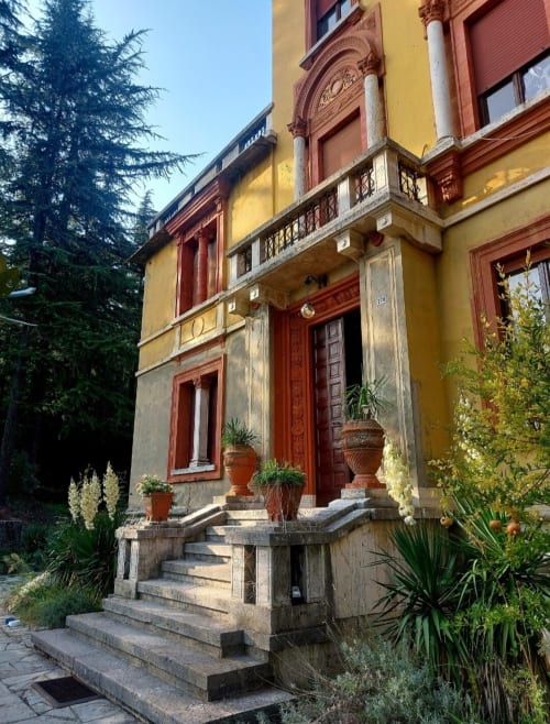 Entrée de villa historique italienne avec façade ocre jaune, portes en bois rouge, escaliers en pierre, jardinières en terre cuite et verdure luxuriante