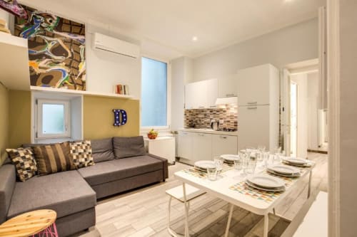 Zona living moderna con divano grigio, tavolo apparecchiato per due, cucina bianca e parete gialla a Roma