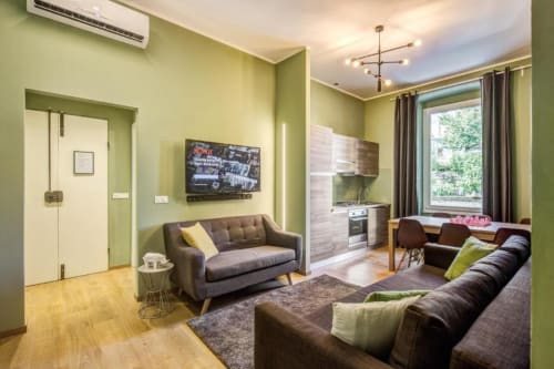 Modern apartment living room with green walls, brown sofa, kitchenette, flat-screen TV, and large window in Roma
