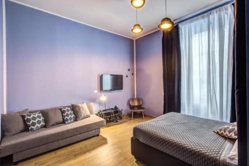 Modern studio apartment in Rome with purple walls, bed, gray sofa, pendant lights, wooden floors, and large windows