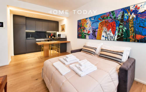 Modern studio apartment in Padova with beige bed, striped pillows, colorful wall art, and black kitchen with wooden bar
