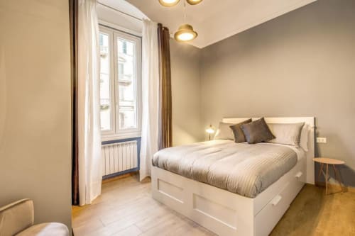 Bright modern bedroom in Roma apartment with white bed, wooden floors, brass pendant lights, and large window with city views