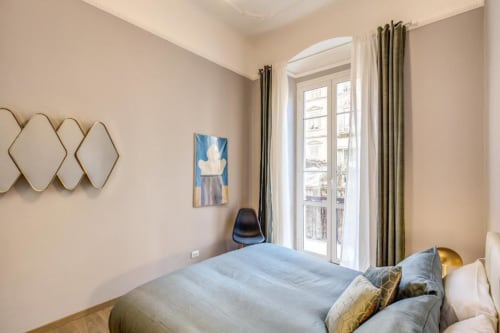 Bright bedroom in Rome apartment with blue bedding, gold pillows, arched mirrors, balcony doors with olive drapes, and wood