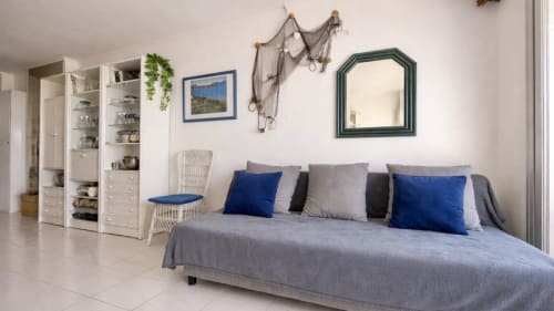 Luminoso apartamento estudio en Holidays Costa Brava con sofá cama gris, cojines azules, unidad de almacenamiento blanca y vistas al mar