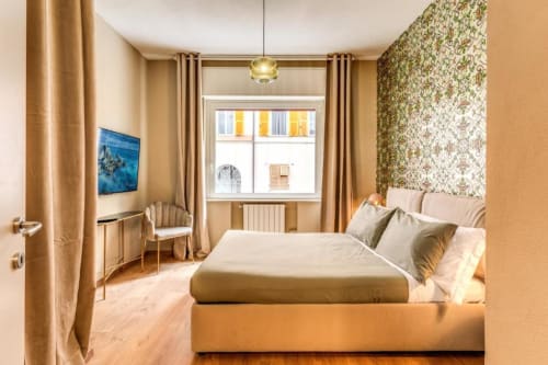 Accogliente camera da letto in appartamento romano con letto matrimoniale, parete con motivi floreali, tende beige, TV e finestra con vista sul giardino