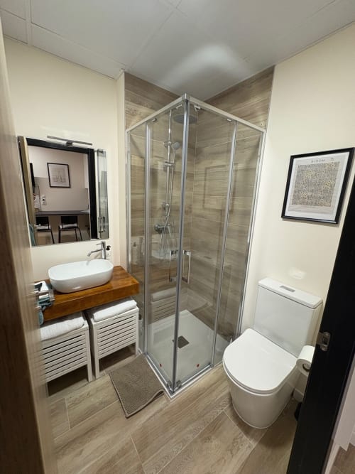 Baño moderno en Be Gonfre Flats Valencia con ducha de cristal, lavabo blanco, inodoro y suelos efecto madera