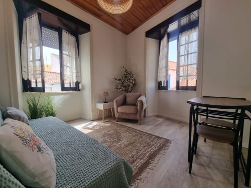 Bright Ericeira hotel room with bed, armchair, wooden ceiling, two windows with ocean views, and dining table