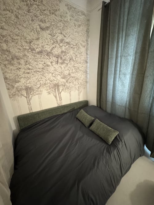 Cozy bedroom at Bouc Bel Air cottage with black bed, green pillows, tree wallpaper, and curtains
