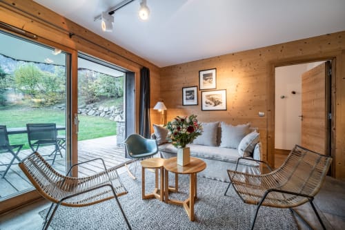 Bright alpine apartment living room with wood paneling, modern seating, mountain views through glass doors, and balcony