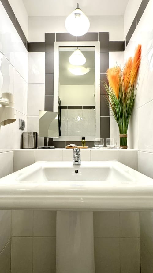 Modernes Waschbecken mit weißem Becken, verchromtem Wasserhahn, großem Spiegel, Pendelleuchte und orangefarbiger Blumendeko im Résidence