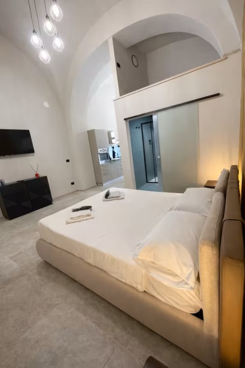 Suite moderna a Bari con letto bianco, soffitto ad arco, luci a sospensione, TV a schermo piatto e accesso a cucinetta