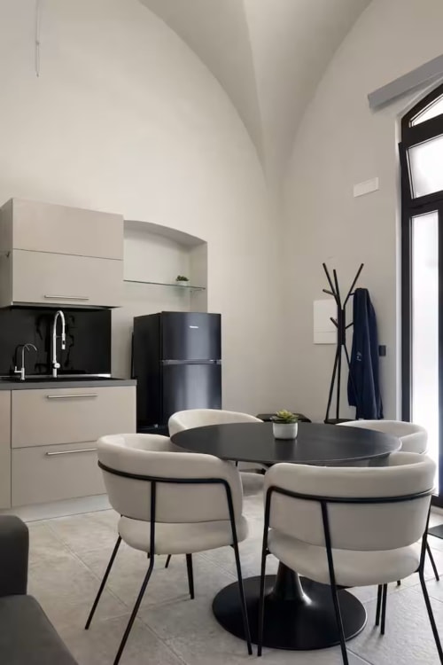 Zona pranzo moderna della Suite 169 a Bari con tavolo rotondo nero, sedie color crema, angolo cottura e pareti curve bianche