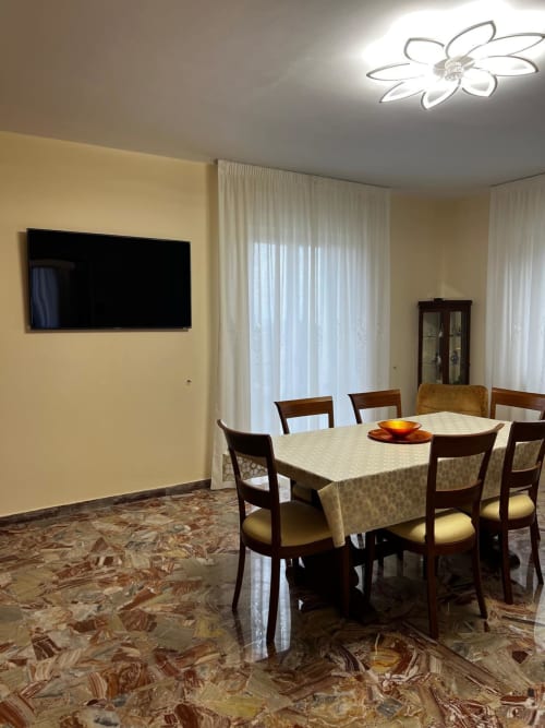 Sala da pranzo moderna con sedie in legno, tavolo, TV e lampada a forma di fiore nel soffitto dell'appartamento Tales of Abruzzo